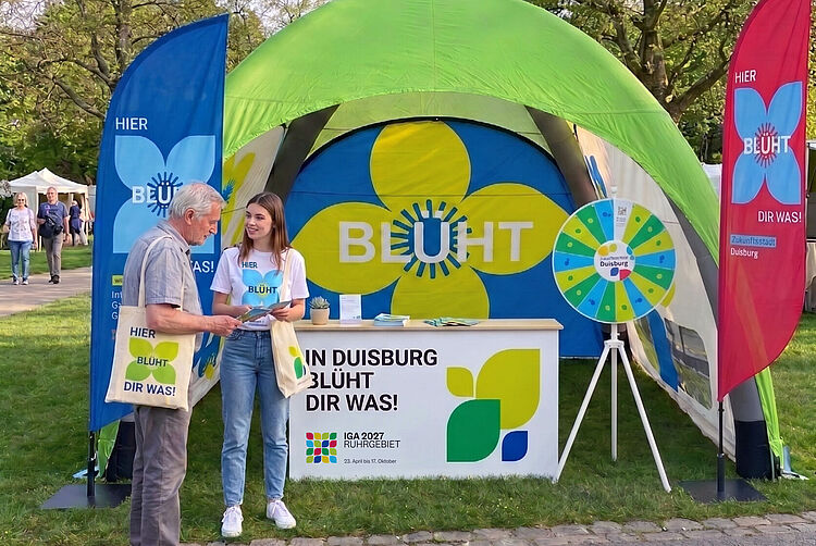 Zelt mit grünem Dach und Werbestand mit gelber Blume und Schriftzug "BLÜHT" sowie zwei Personen, die sich unterhalten, umgeben von blauen und roten Fahnen mit demselben Schriftzug