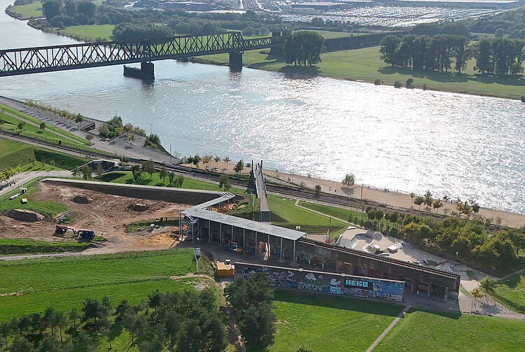 Luftaufnahme eines Flusses mit einer Eisenbahnbrücke, angrenzenden Grünflächen und einem Baugelände mit Baugerüsten des Skywalks.