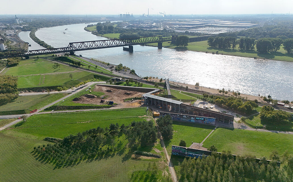 Luftaufnahme eines Flusses mit einer Eisenbahnbrücke, angrenzenden Grünflächen und einem Baugelände mit Baugerüsten des Skywalks.