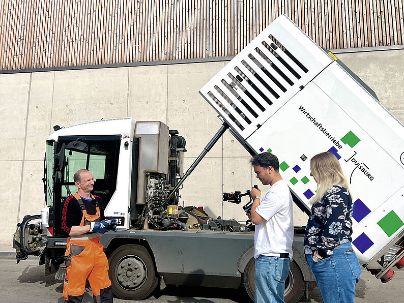 Zwei Personen filmen einen Mann in orangefarbener Arbeitskleidung vor einem geöffneten Nutzfahrzeug mit Aufschrift 'Wirtschaftsbetriebe Duisburg'