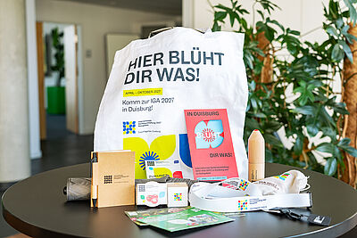 Weiße Stofftasche mit Aufdruck 'HIER BLÜHT DIR WAS!' und verschiedenen Werbeartikeln der IGA 2027 auf einem runden Tisch in einem Innenraum