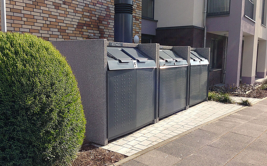 Drei große graue Müllgroßbehälter mit Klappdeckeln stehen sauber nebeneinander auf einem gepflasterten Gehweg vor einem Wohnhaus
