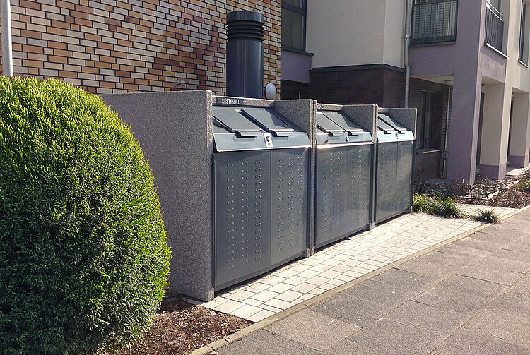 Drei große graue Müllgroßbehälter mit Klappdeckeln stehen sauber nebeneinander auf einem gepflasterten Gehweg vor einem Wohnhaus