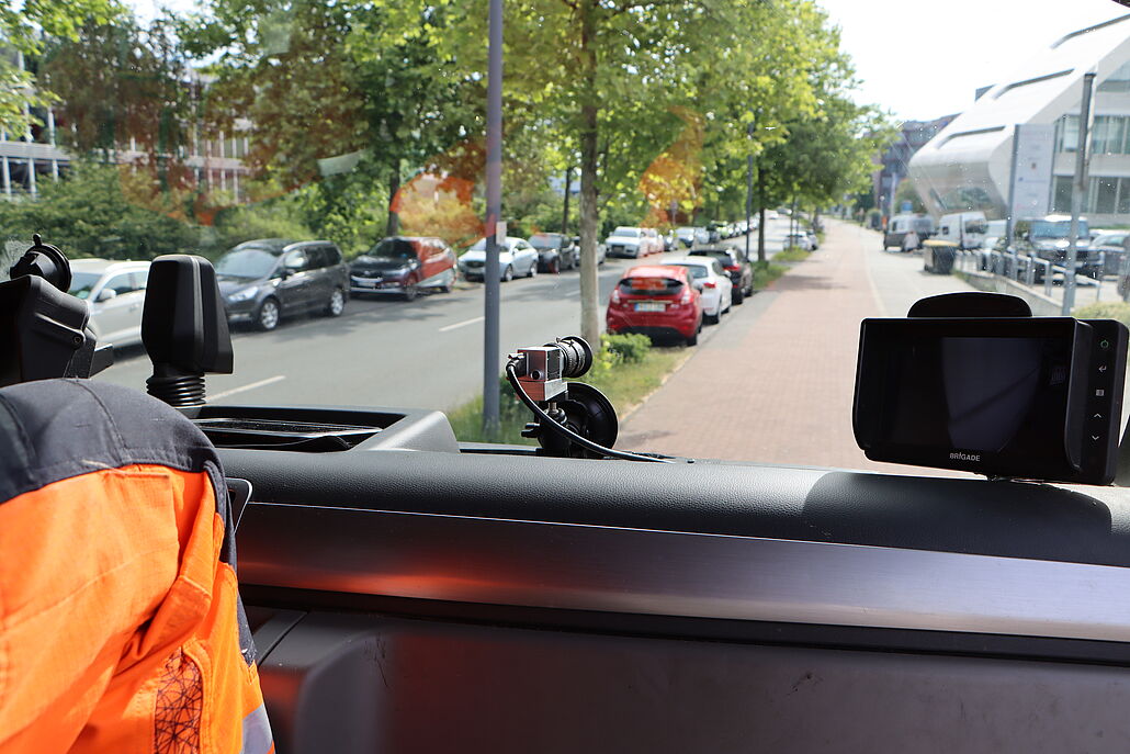 Blick aus einem Fahrzeug auf eine Straße mit geparkten Autos und Bäumen, links ein orangefarbener Arbeitsanzug sichtbar