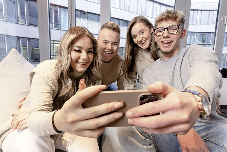 Vier junge Erwachsene sitzen auf einem Sofa und machen gemeinsam ein Selfie mit einem Smartphone