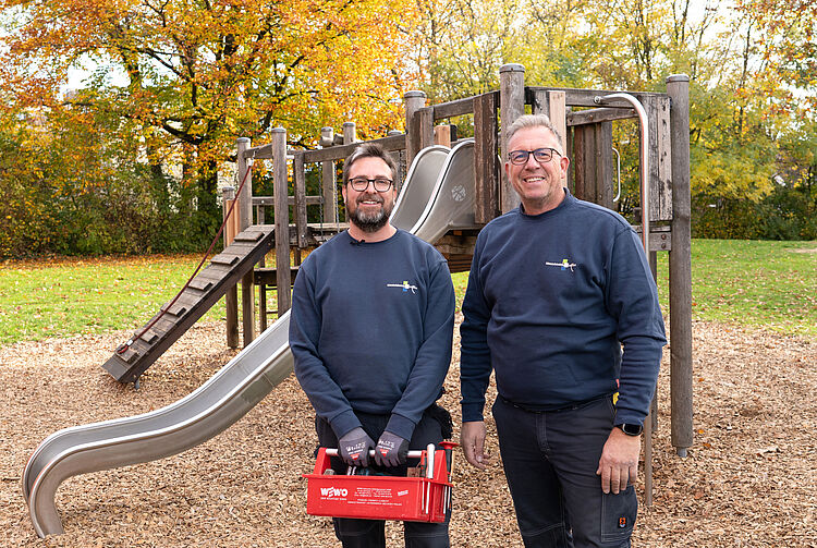 Zwei Männer in blauen Pullovern stehen vor einem Spielplatz mit Rutsche und Klettergerüst, einer hält eine rote Werkzeugkiste