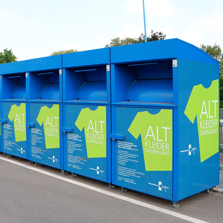Sechs blaue Altkleidercontainer mit grüner T-Shirt-Grafik und der Aufschrift 'ALT KLEIDER SAMMLUNG' stehen auf einem Parkplatz.