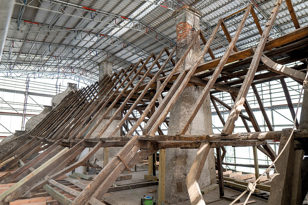 Innenansicht einer Baustelle mit freiliegenden, alten Holzbalken und einem gemauerten Schornstein unter einem Gerüstdach