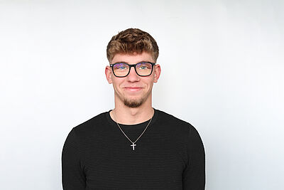 Junger Mann mit kurzen braunen Haaren trägt schwarzen Pullover und silberne Halskette mit Kreuzanhänger vor hellem Hintergrund.