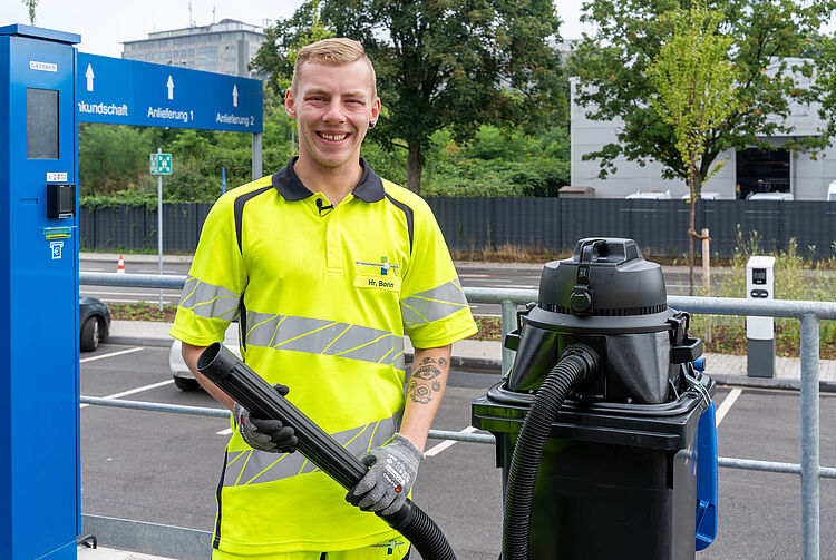 Person in gelb-schwarzer Warnschutzkleidung steht neben schwarzem Müllcontainer mit aufgesetztem Industriesauger und hält Schlauch in der Hand.