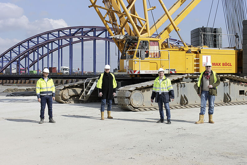 Vier Personen in Sicherheitskleidung stehen vor einem großen gelben Kran auf einer Baustelle mit einer Stahlbogenbrücke im Hintergrund