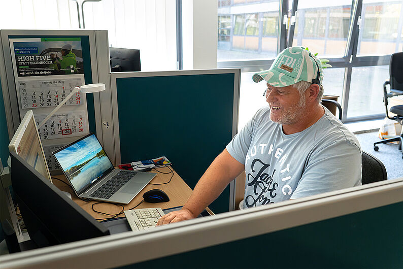 Mitarbeiter im Kundencenter der WBD sitzt lächelnd an einem Schreibtisch mit Laptop und mehreren Monitoren in einem hellen Büro mit großen Fenstern