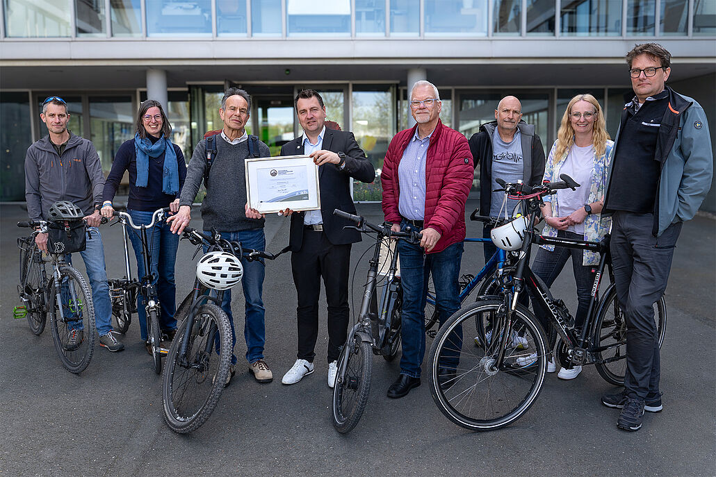 Acht Mitarbeitende der WBD stehen mit Fahrrädern vor einem Gebäude, eine Person hält eine gerahmte Auszeichnung