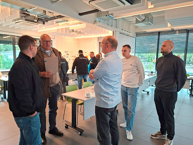 Mehrere Männer stehen in einem modernen, hellen Raum mit großen Fenstern und weißen Tischen, einige unterhalten sich.