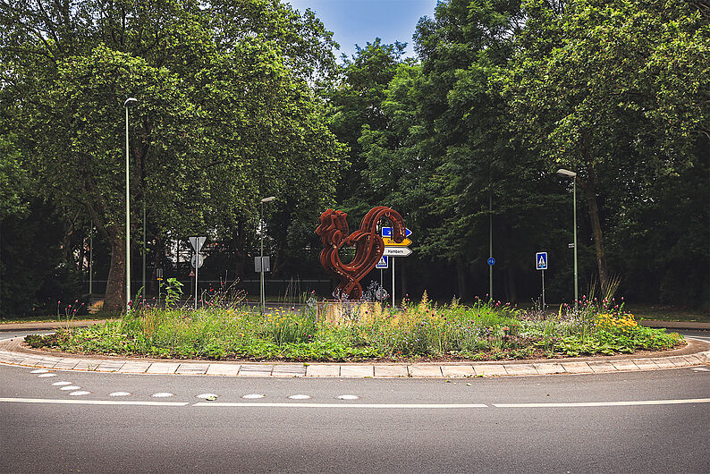 Kreisverkehr mit bepflanzter Mittelinsel und einer großen roten Skulptur in Form eines Hahns, umgeben von Bäumen und Verkehrszeichen